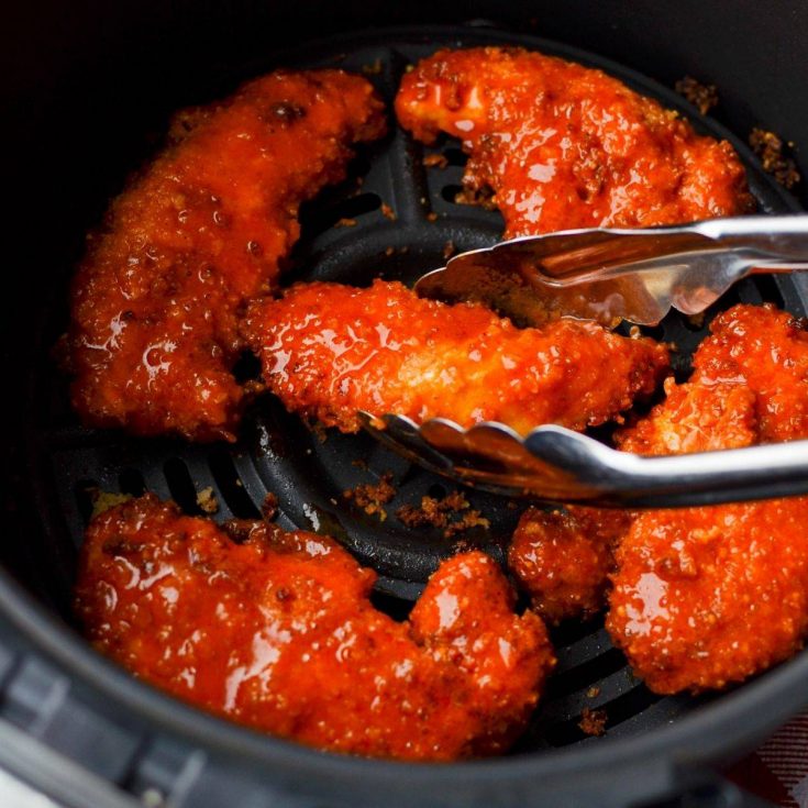 An air fryer basket filled with buffalo chicken tenders, and a pair of tongs taking them out.