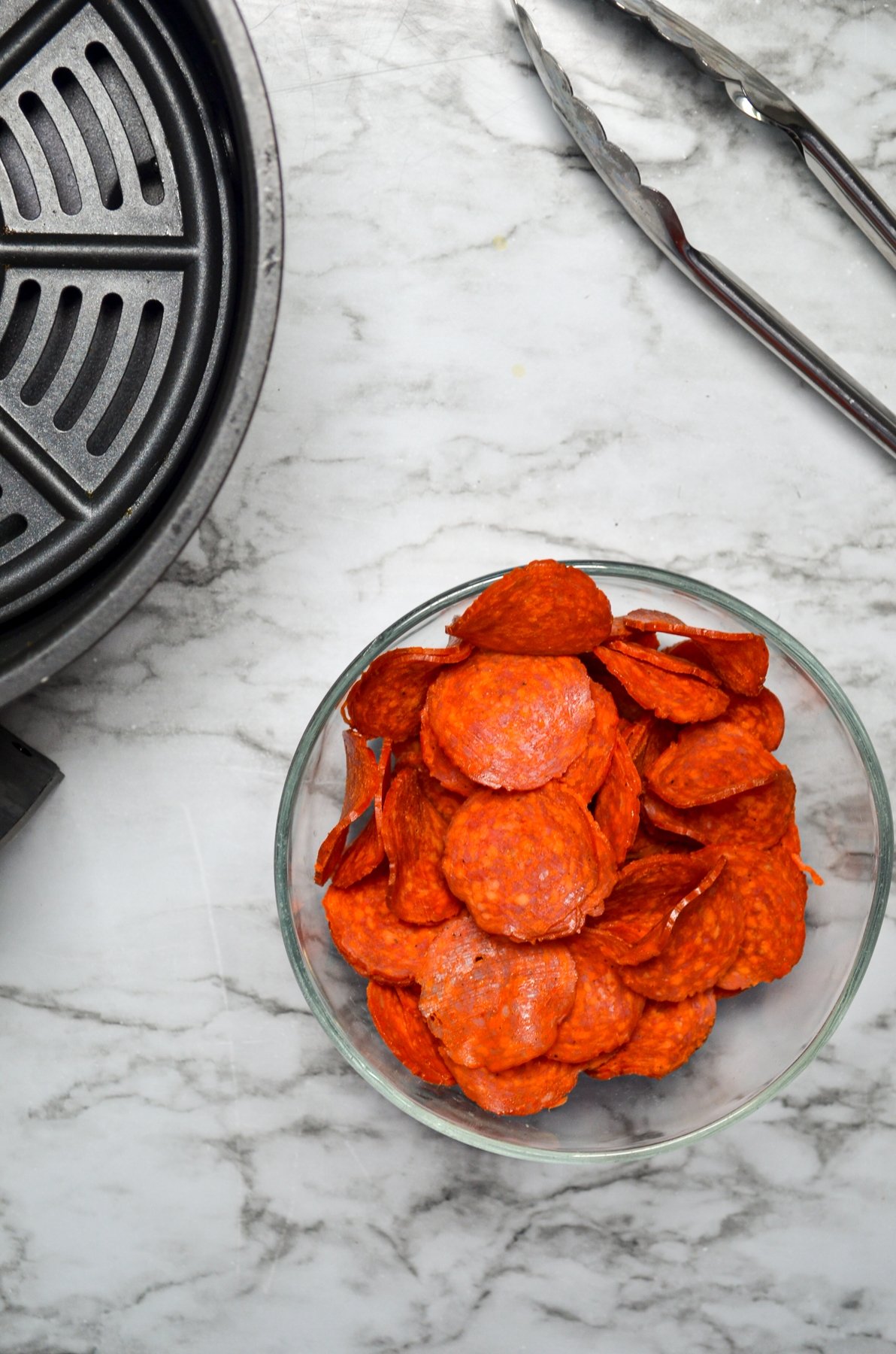 A bowl of pepperoni, with an air fryer basket and a pair of tongs on the side.