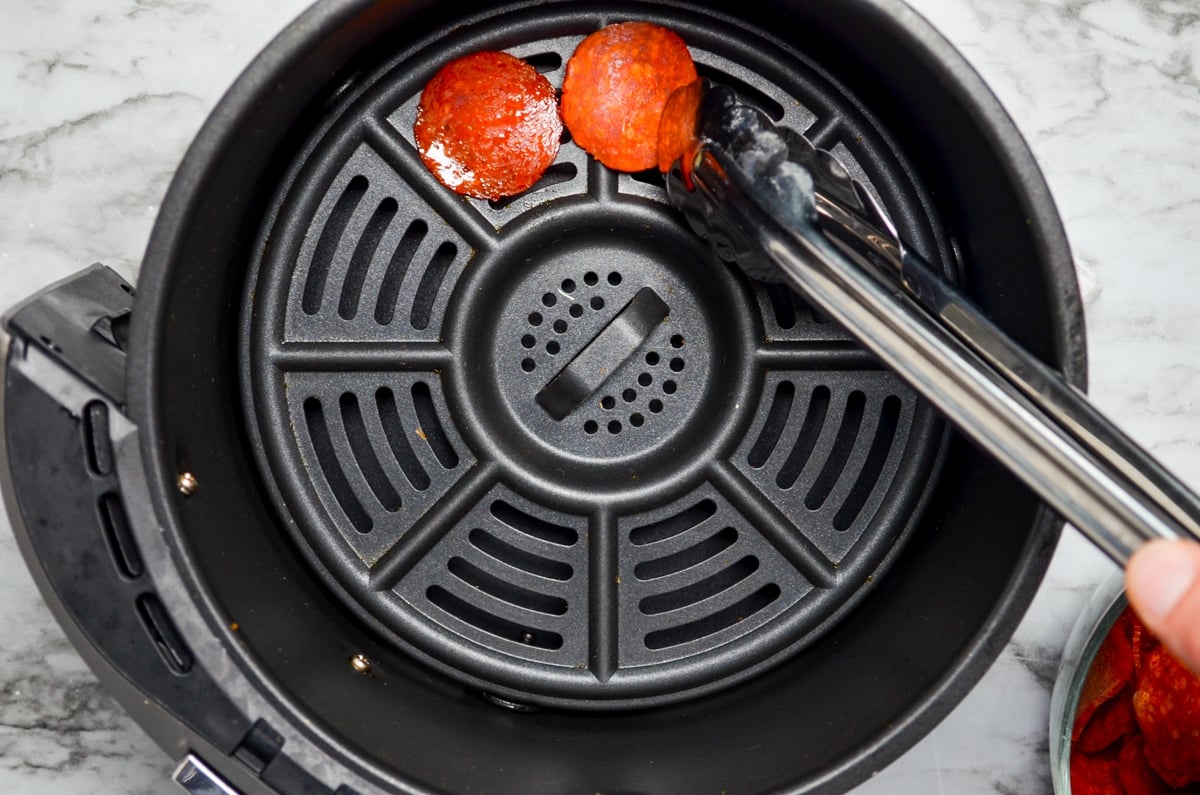 A set of tongs placing pepperoni slices in the basket of an air fryer.