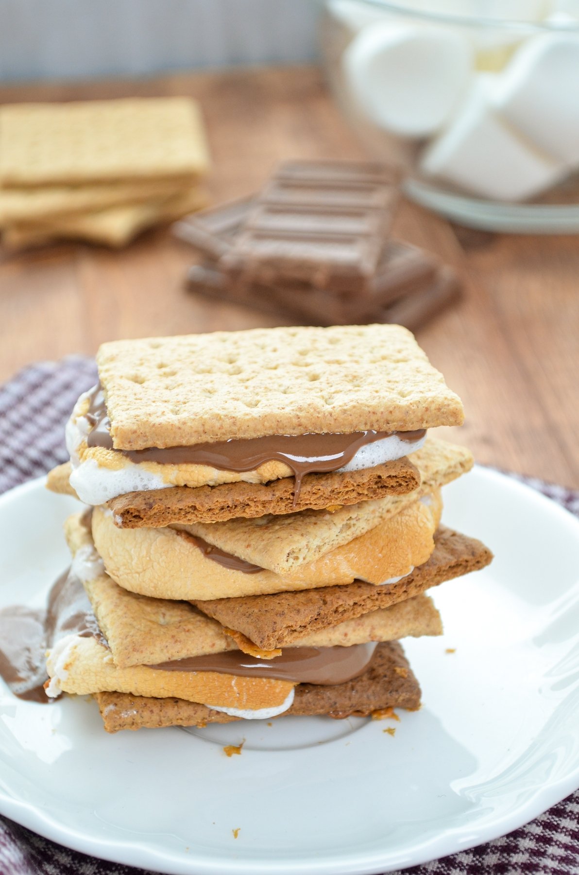 A stack of three s'mores with chocolate, marshmallows, and graham crackers in the background.
