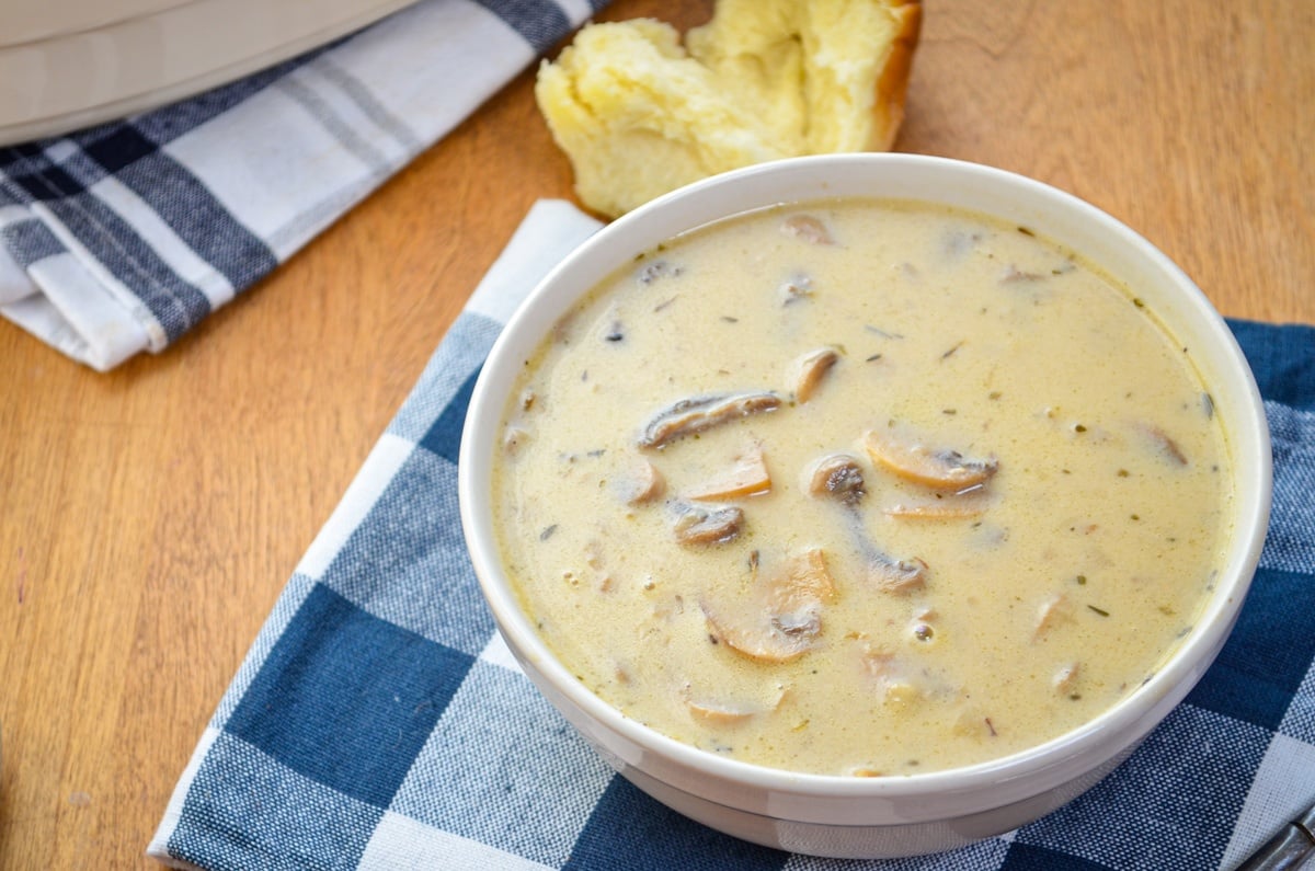 A bowl of cream of mushroom soup with a torn roll in the background.