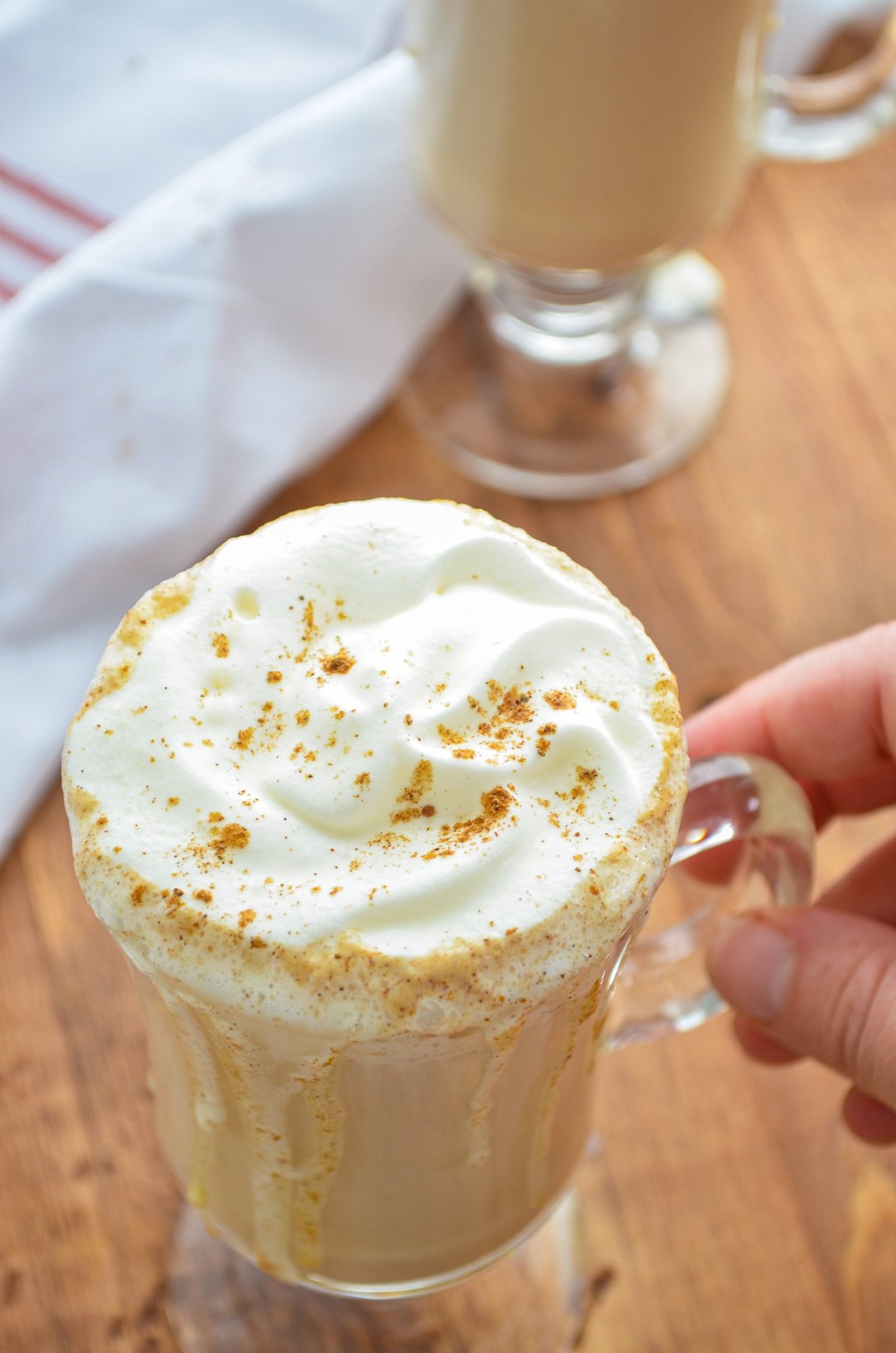 A glass mug filled with a flavored coffee and whipped cream is being lifted by a hand.
