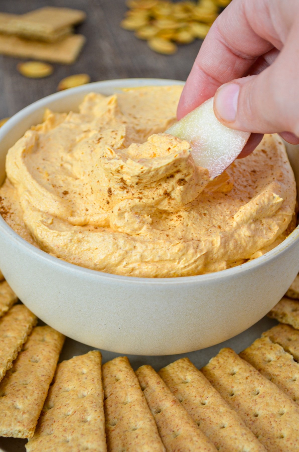 A bowl of pumpkin fluff dip, surrounded by graham crackers. A hand dips an apple slice into the orange colored dip.