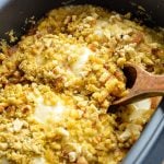 A wooden spoon taking a swiss cheese and stuffing topped chicken breast from a slow cooker.