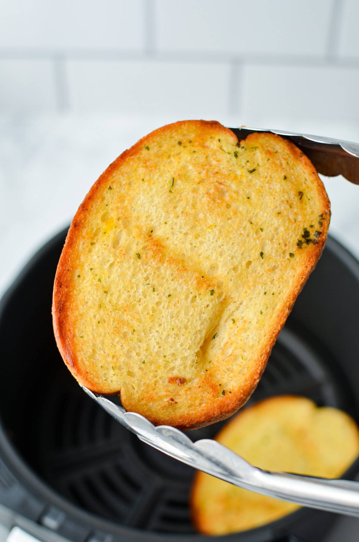 Using a pair of tongs to hold up a toasted piece of frozen garlic bread.