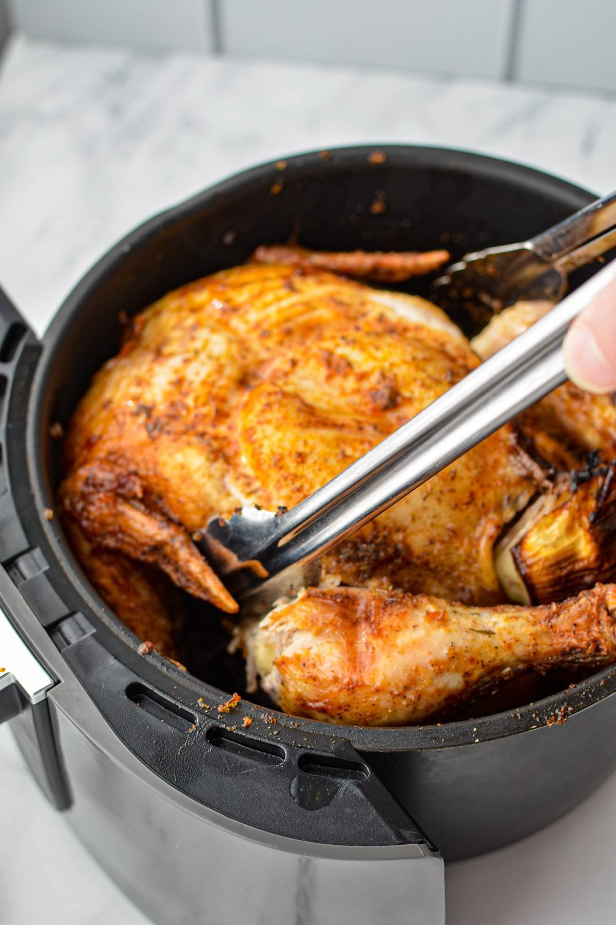 Using tongs to flip a whole chicken in an air fryer basket.