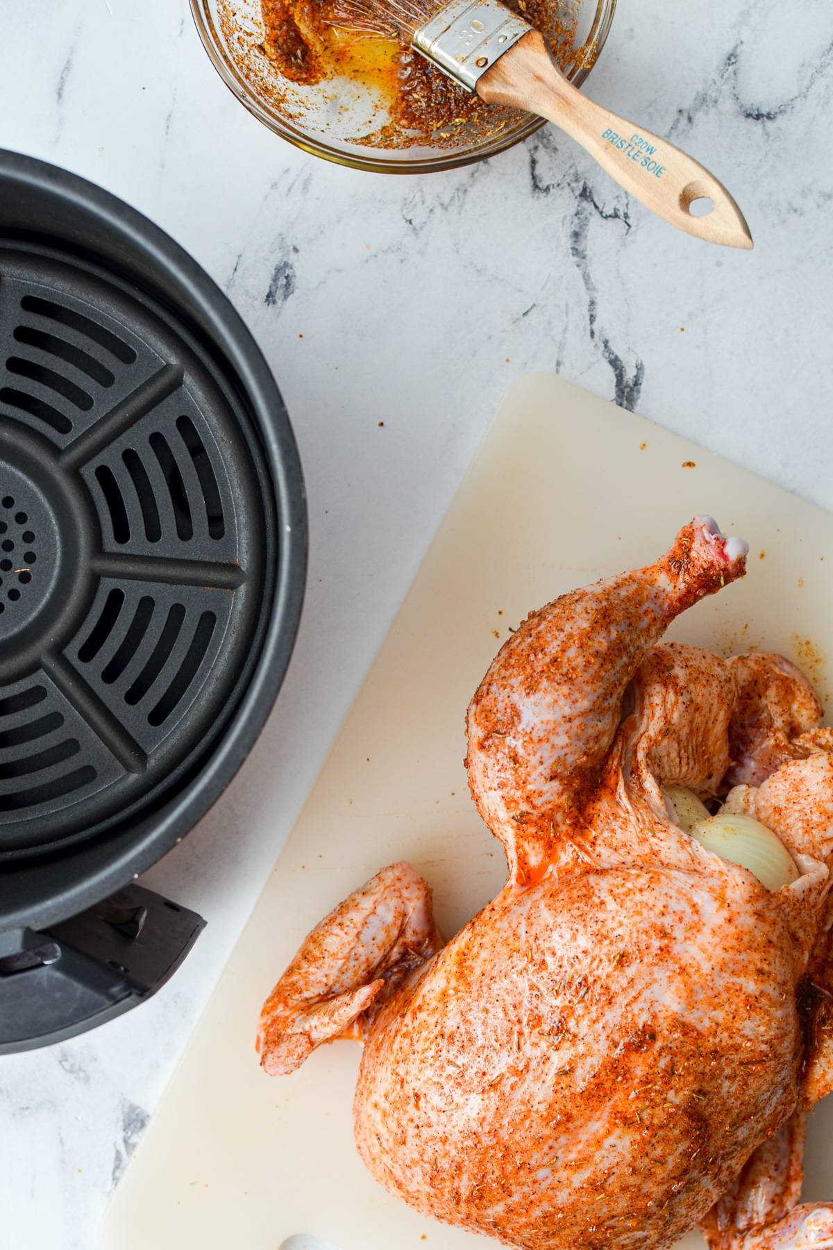 A whole chicken brushed with a spice rub, with an air fryer basket on the side.