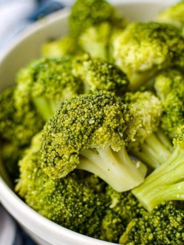 A bowl of cooked broccoli, resting on a blue check napkin.