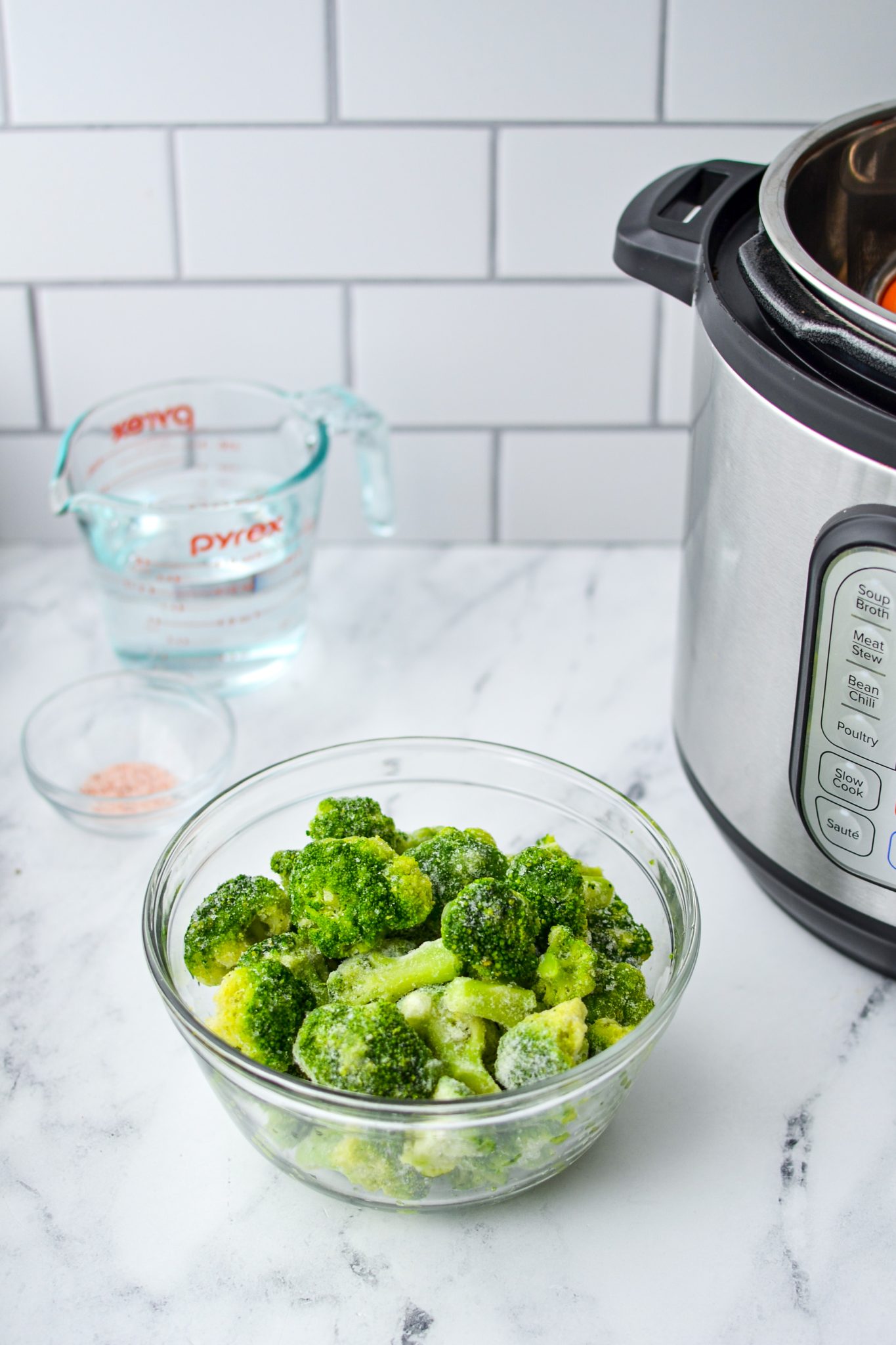 A bowl of frozen broccoli on a white marble countertop. A measuring cup of water, some salt, and an Instant Pot in the background.