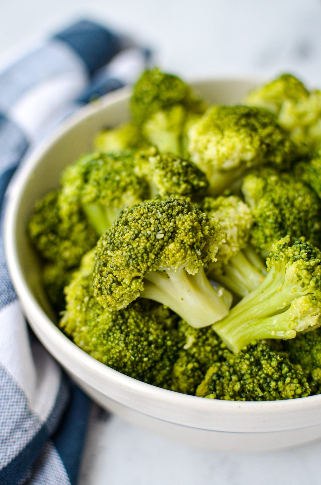 A bowl of cooked broccoli, resting on a blue check napkin.