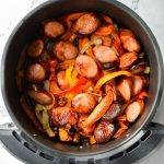 An air fryer basket, filled with cooked sausage, peppers, and onions.