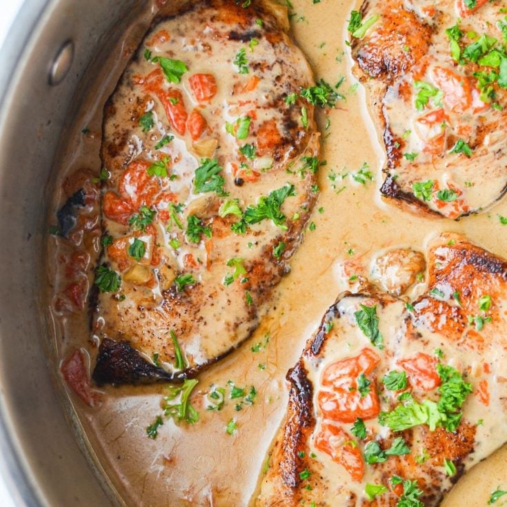 A skillet filled with chicken breasts coated in a red pepper cream sauce, and garnished with parsley.