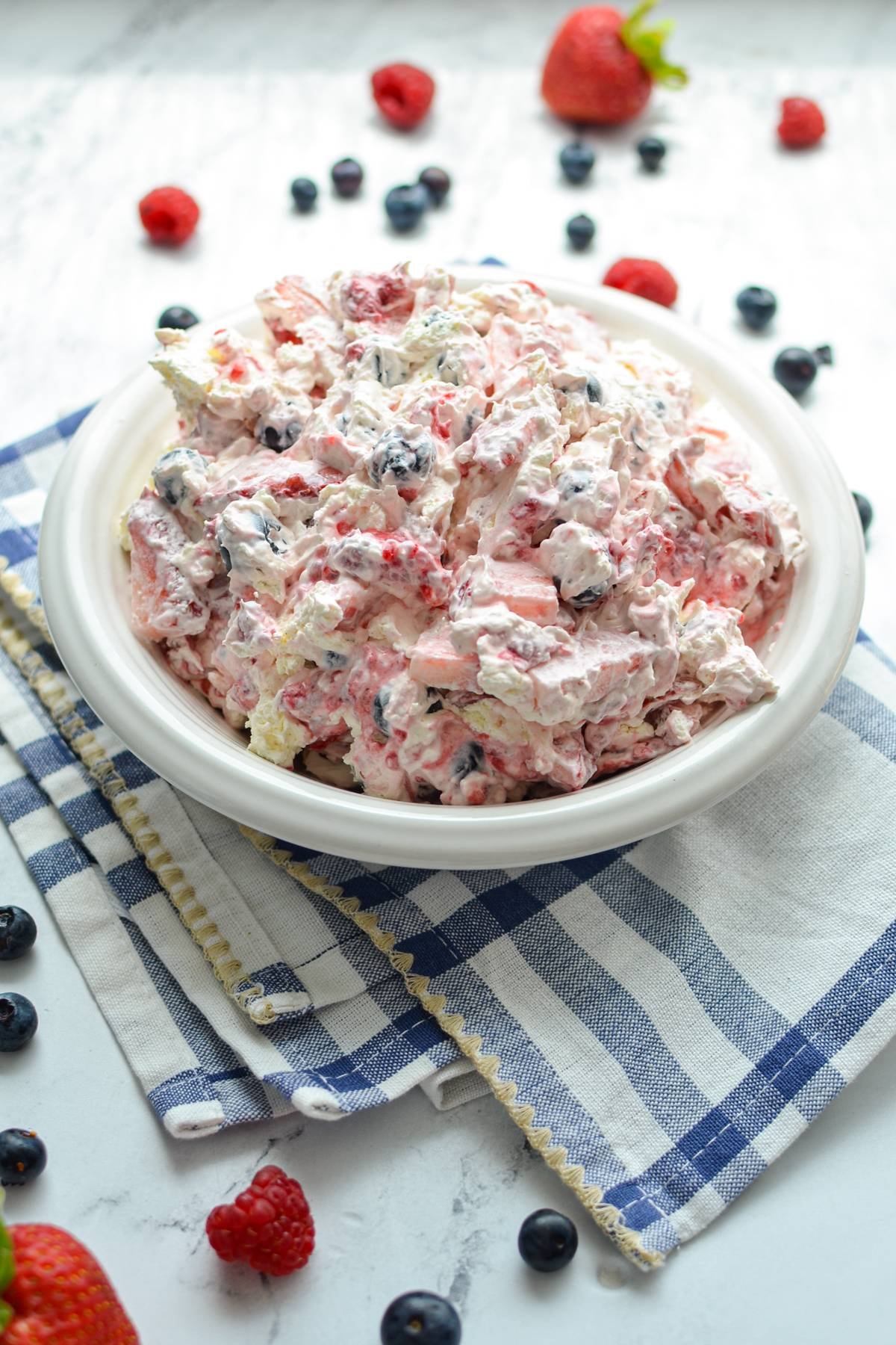 A bowl of red white and blue cheesecake salad, in a bowl resting on top of a blue and white kitchen towel.