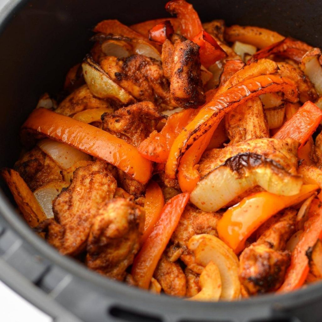 An air fryer basket with chicken, peppers, and onions inside.