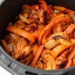 An air fryer basket with chicken, peppers, and onions inside.