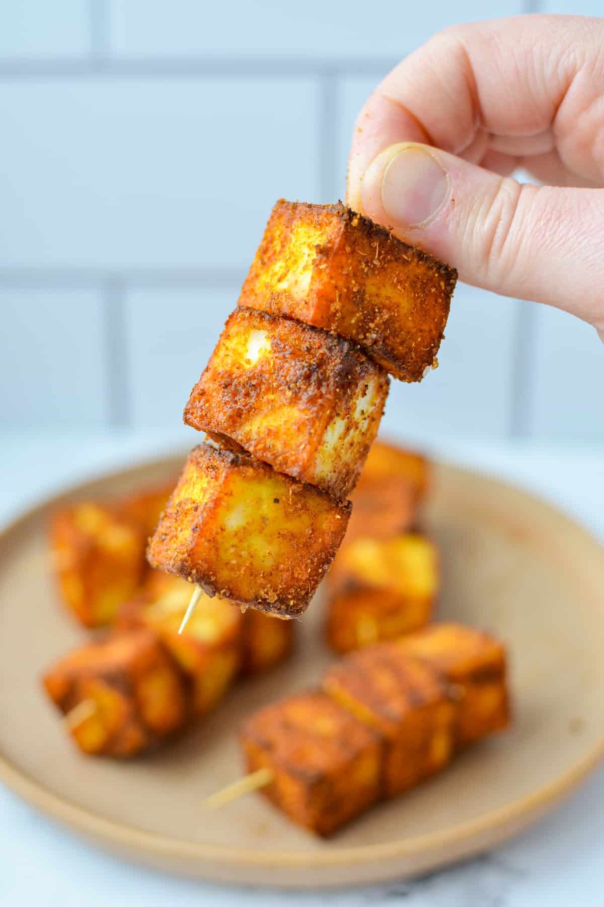 A hand holding a skewer with three cubes of seasoned paneer speared onto it.