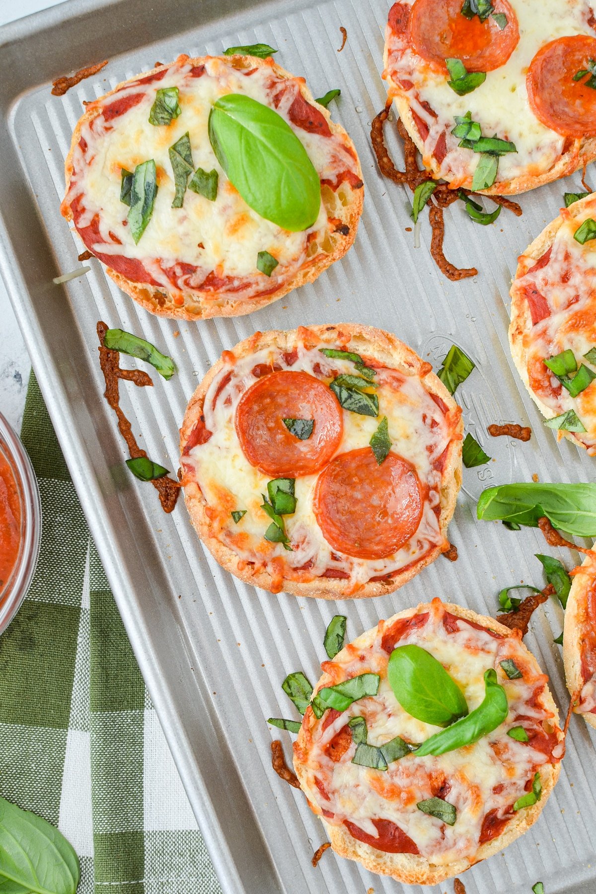 A baking sheet with English muffin pizzas topped with pepperoni and basil.