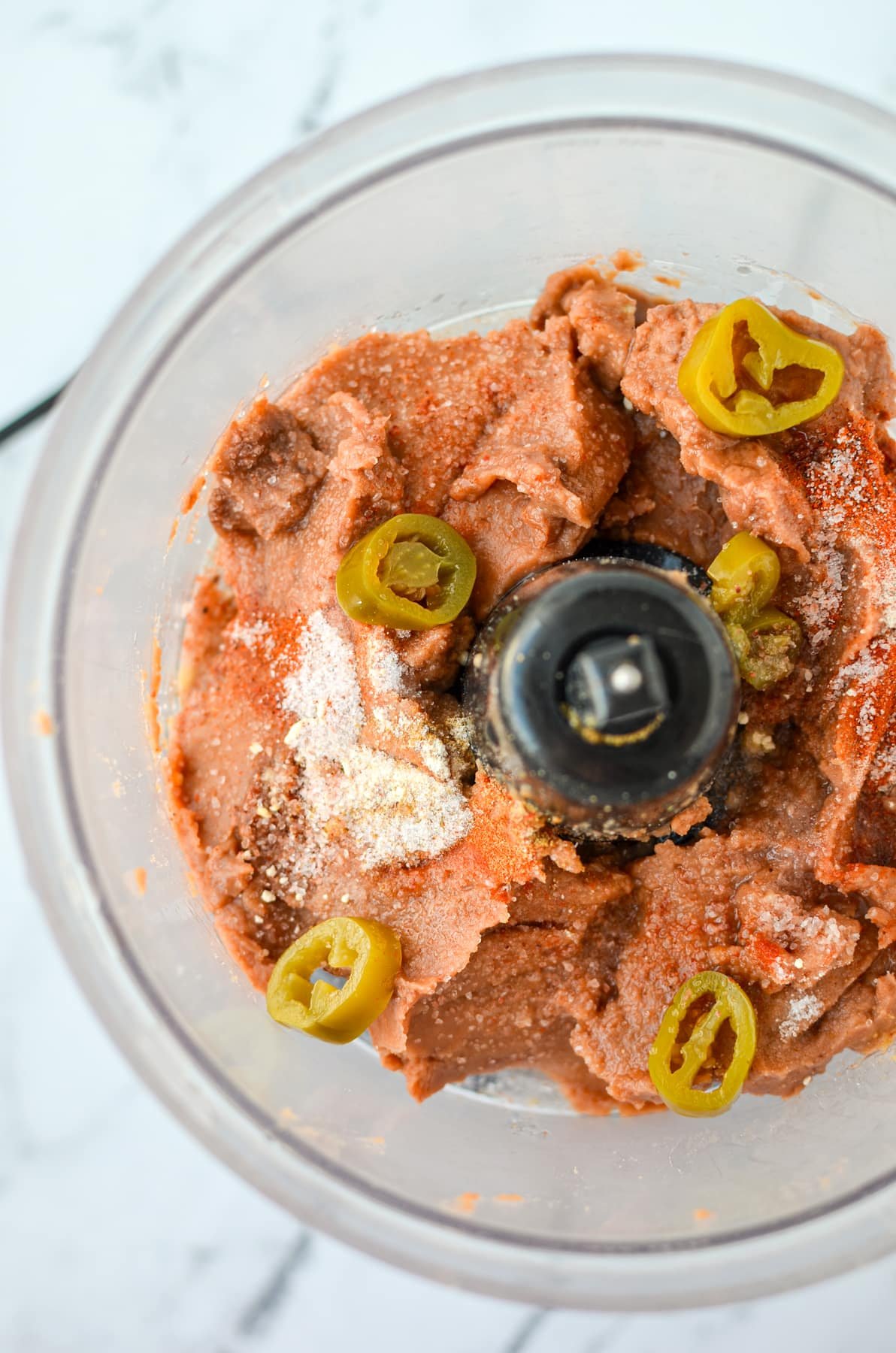 A food processor with refried beans, jalapeno slices, and seasonings.