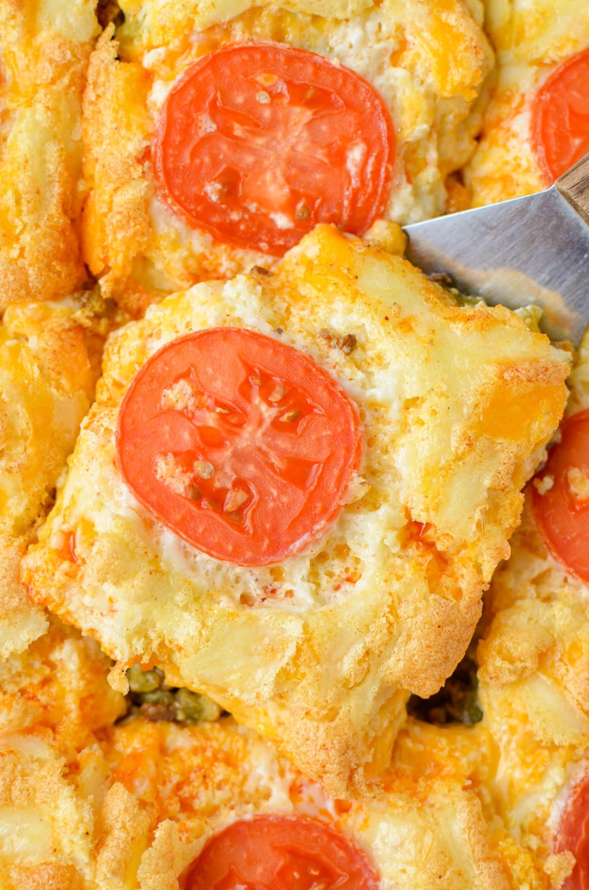 A spatula lifting a cheesy casserole from a dish topped with fresh tomato slices. 