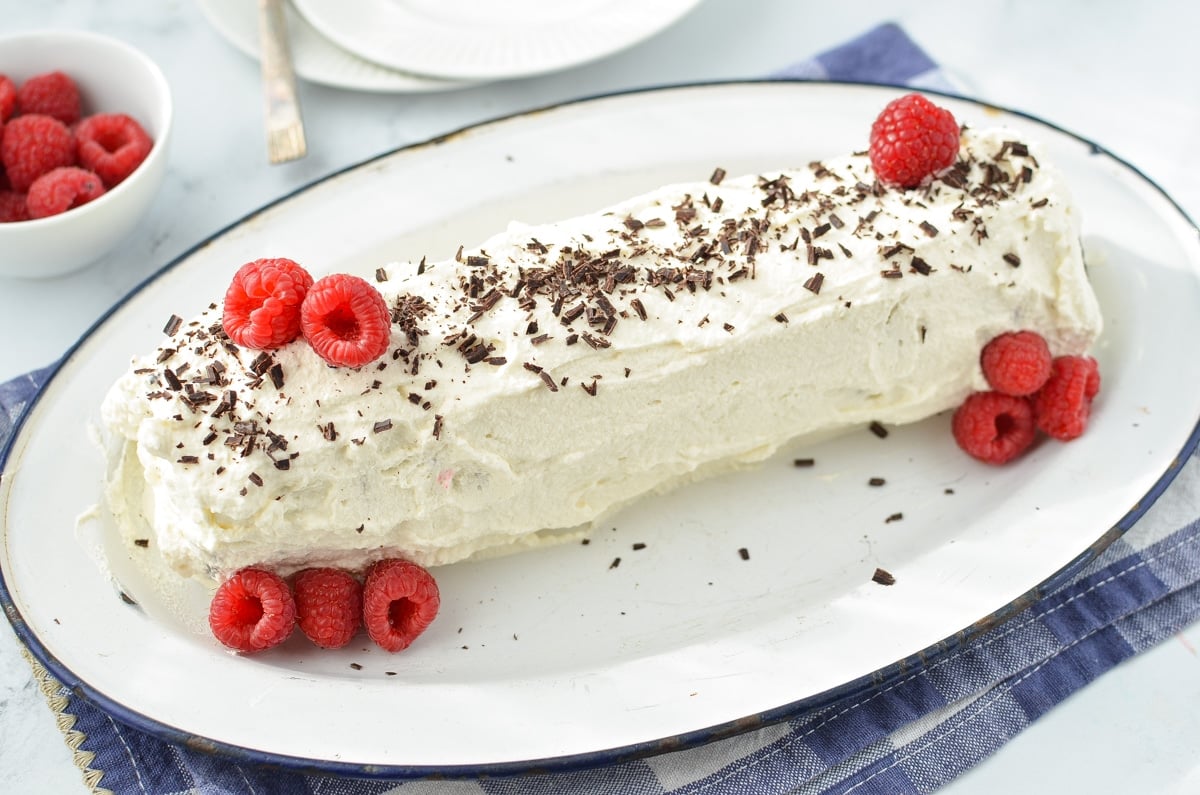 A whipped cream covered log cake, garnished with chocolate shavings and fresh raspberries.