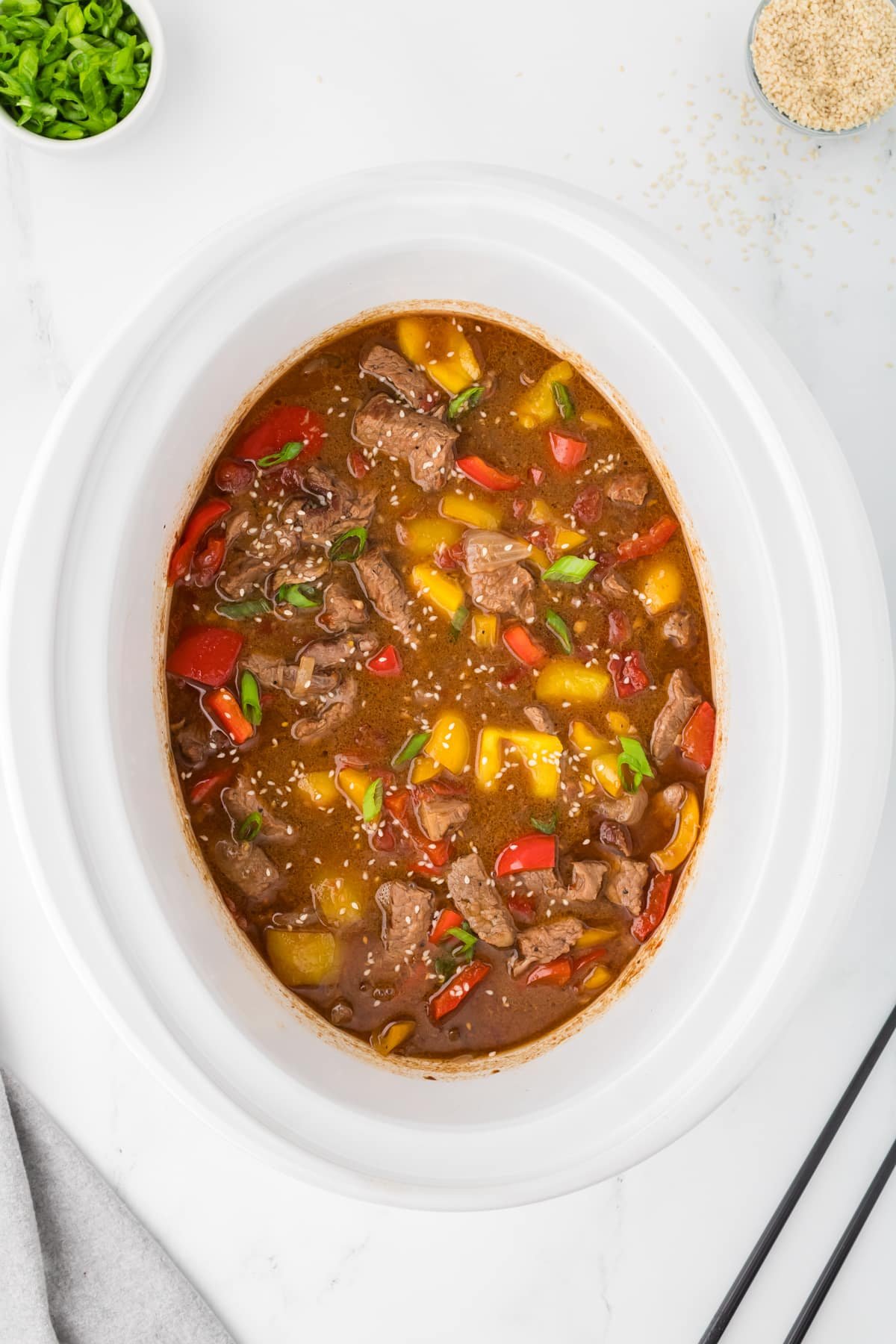 A slow cooker filled with a saucy pepper steak and garnished with sesame seeds.