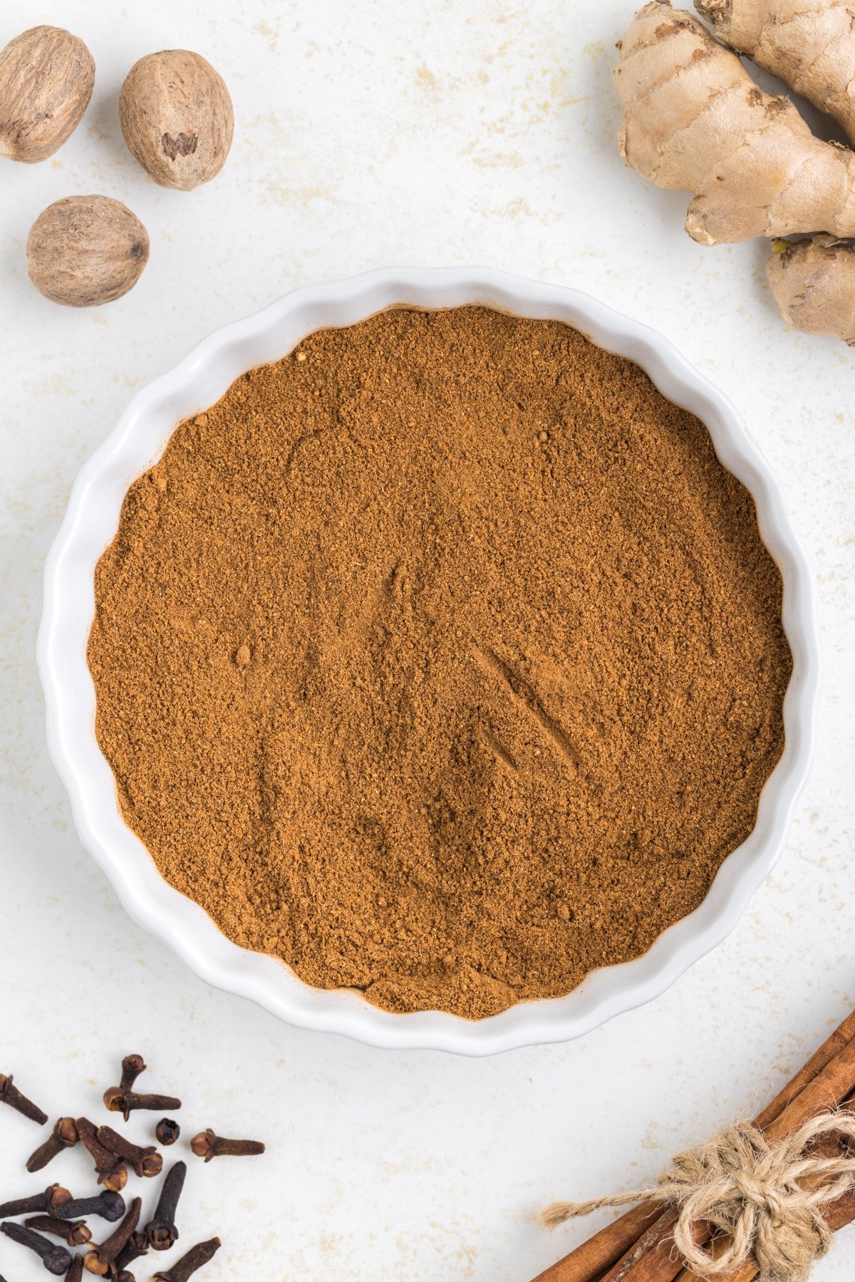 A dish filled with a homemade spice blend, surrounded by ginger root, nutmeg, cloves, and cinnamon.