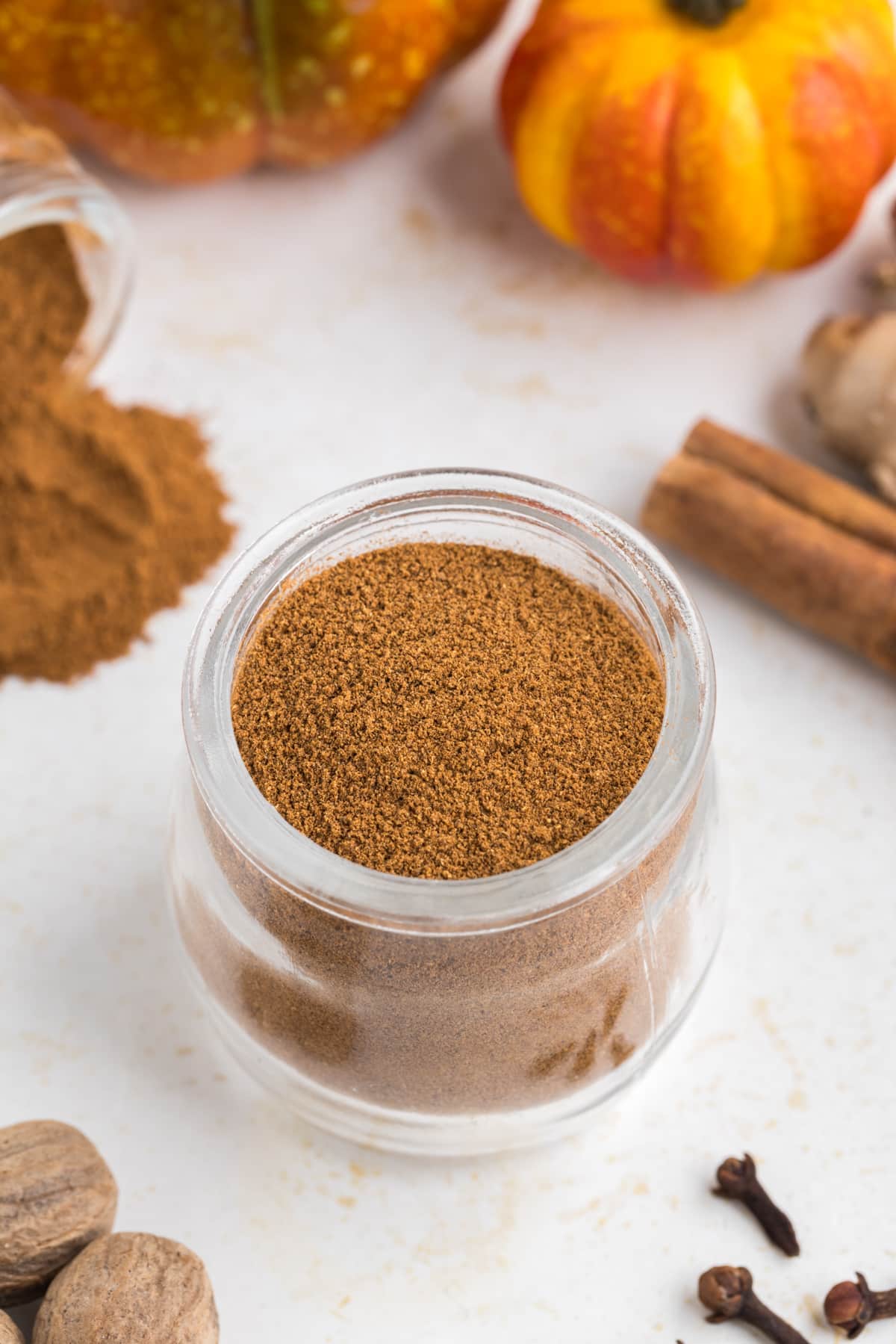 A small jar of pumpkin spice, surrounded by whole spices and mini pumpkins.