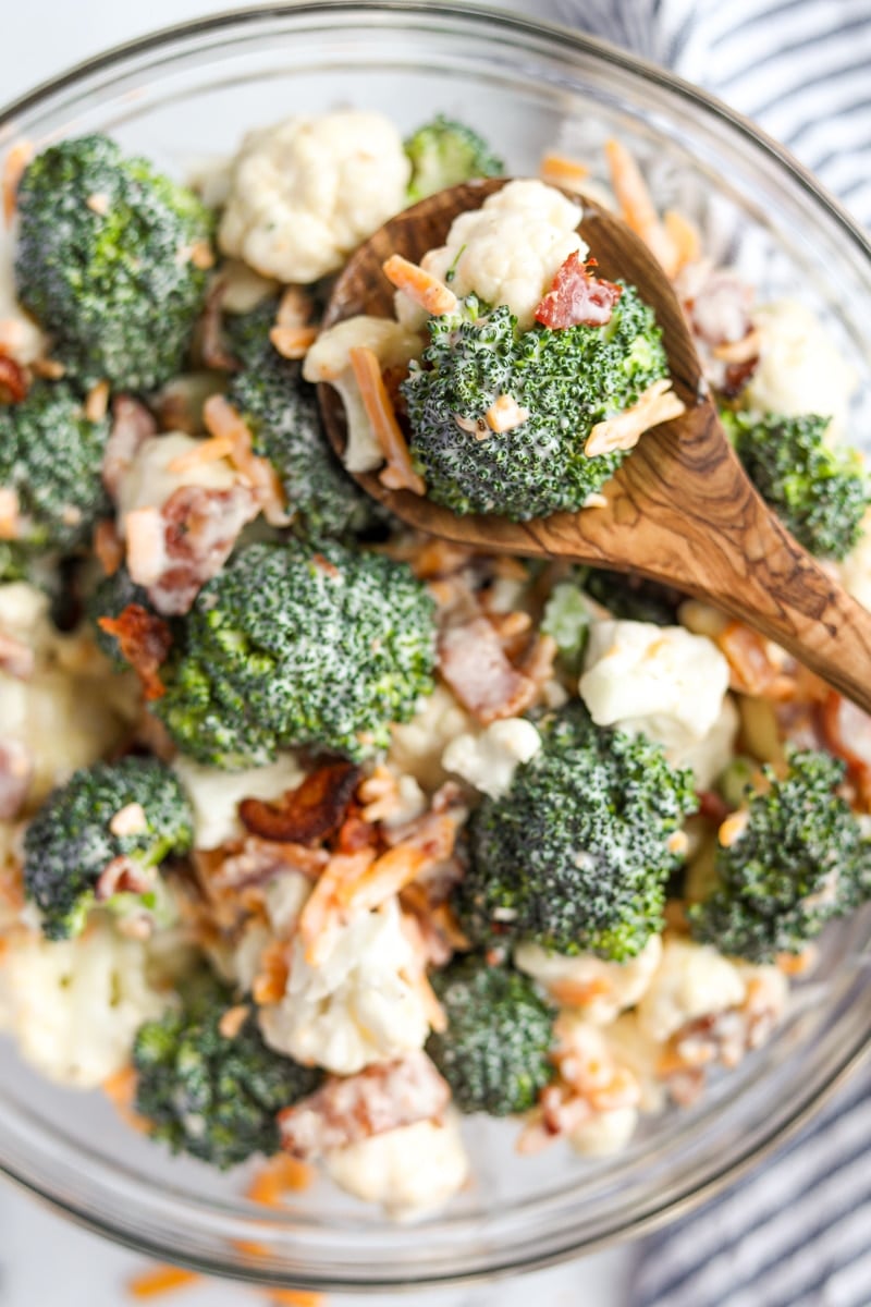 A bowl of broccoli salad with a wooden spoon removing a portion.