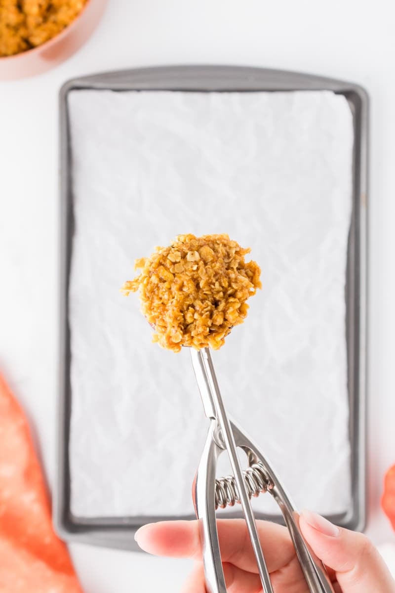 A cookie scoop with an oatmeal mixture hovering over a parchment lined baking sheet.