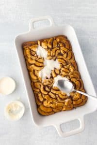 Spreading glaze on a baking dish with baked cinnamon roll pieces.