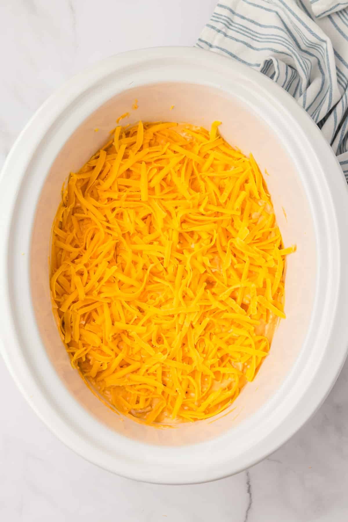 A white crock pot with a cheesy dip inside, topped with shredded cheddar cheese.