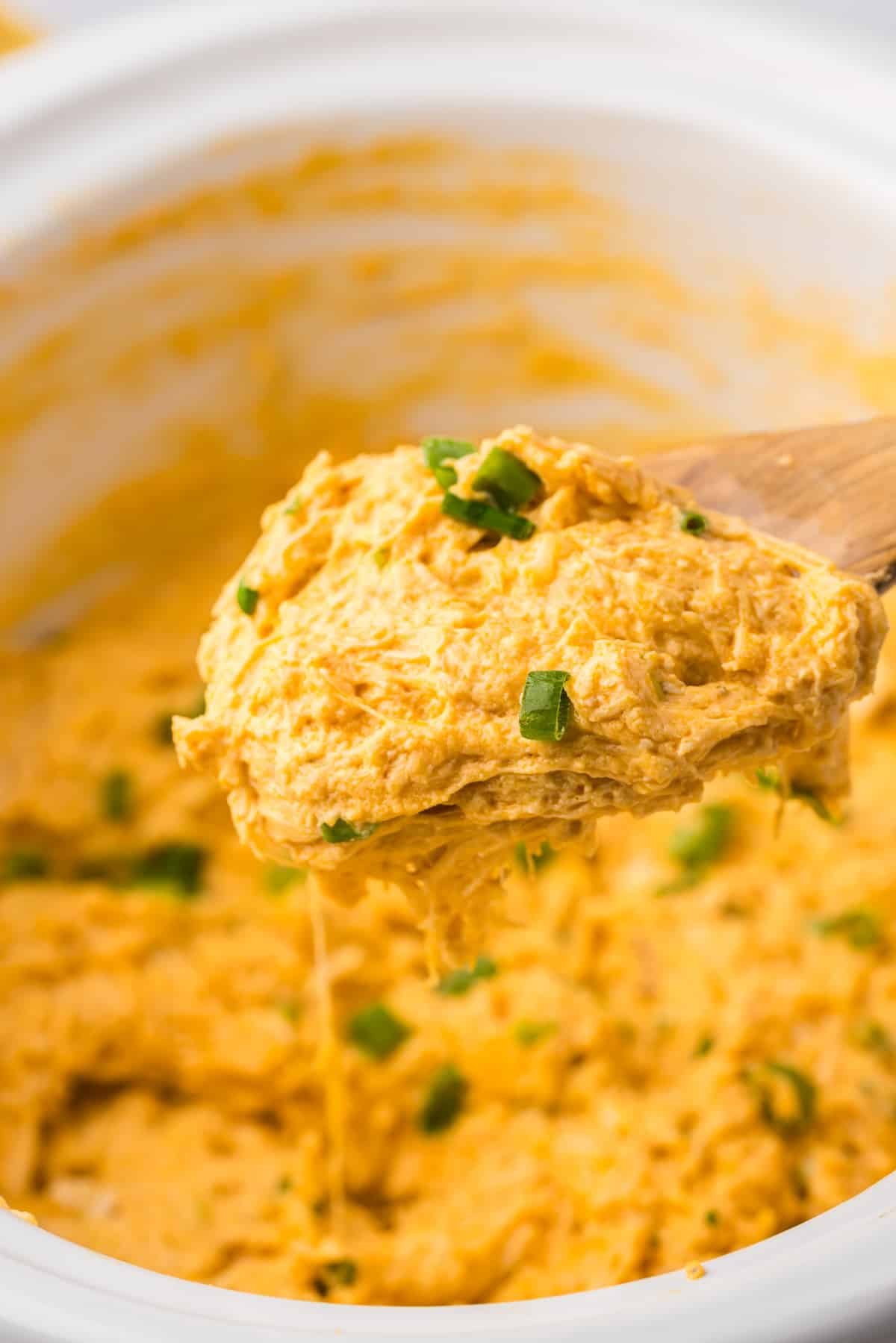 A wooden spoon taking a small amount of chicken dip from a white slow cooker.