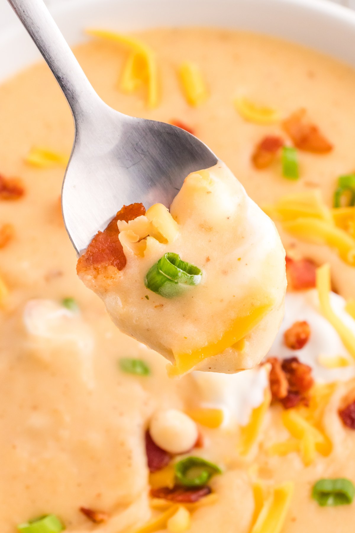 A spoon with cheesy potato soup on it, garnished with bacon and green onions.