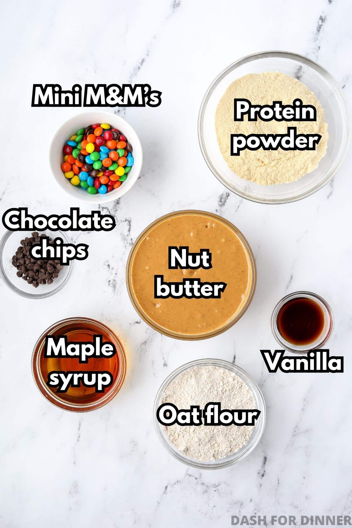 An overhead view of different bowls with different ingredients, including: nut butter, protein powder, mini M&m's, chocolate chips, maple syrup, etc.