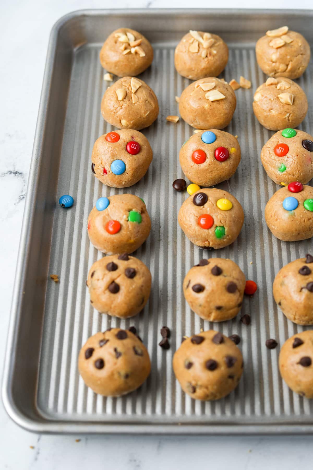 A baking tray with cookie dough balls, featuring chocolate chip, m&m, and chopped peanuts.