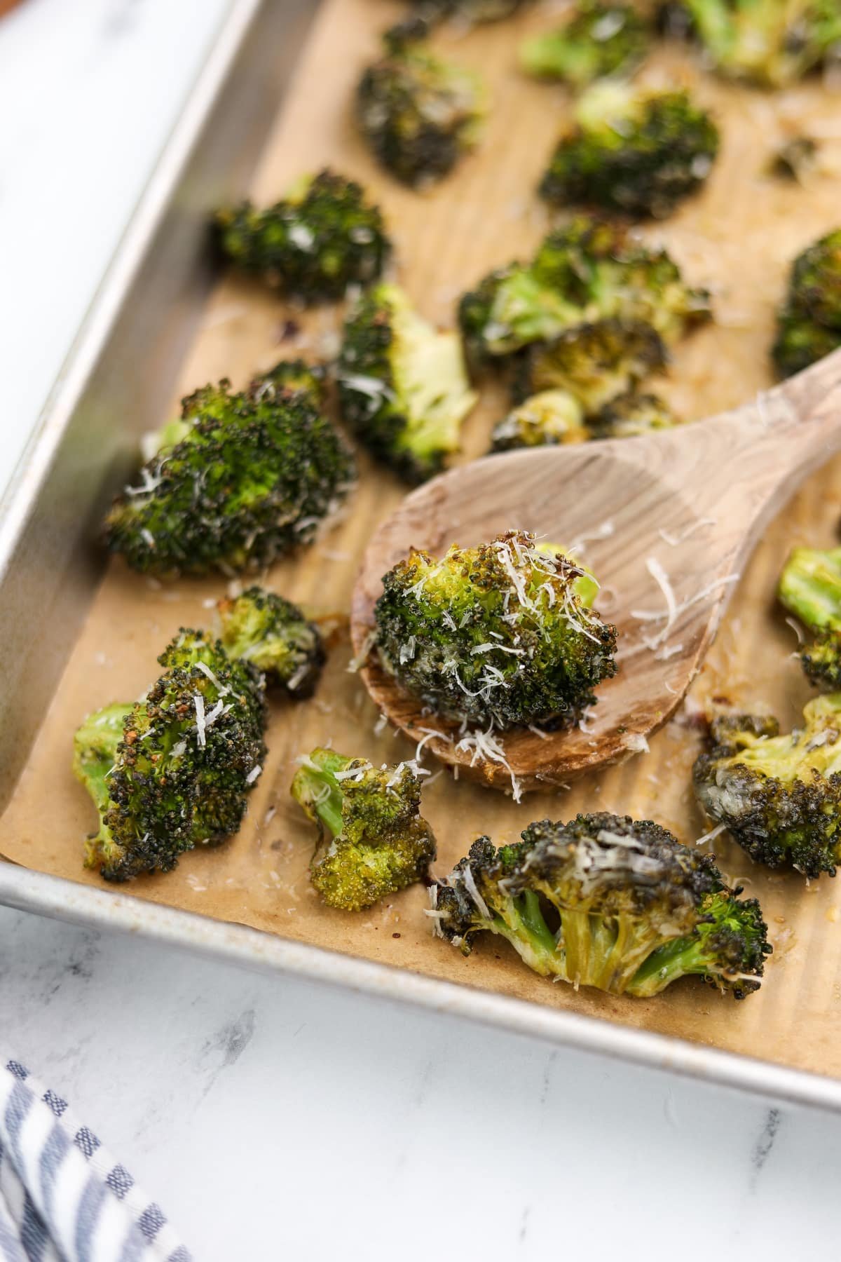 A wooden spoon taking a portion of roasted broccoli from a baking sheet.