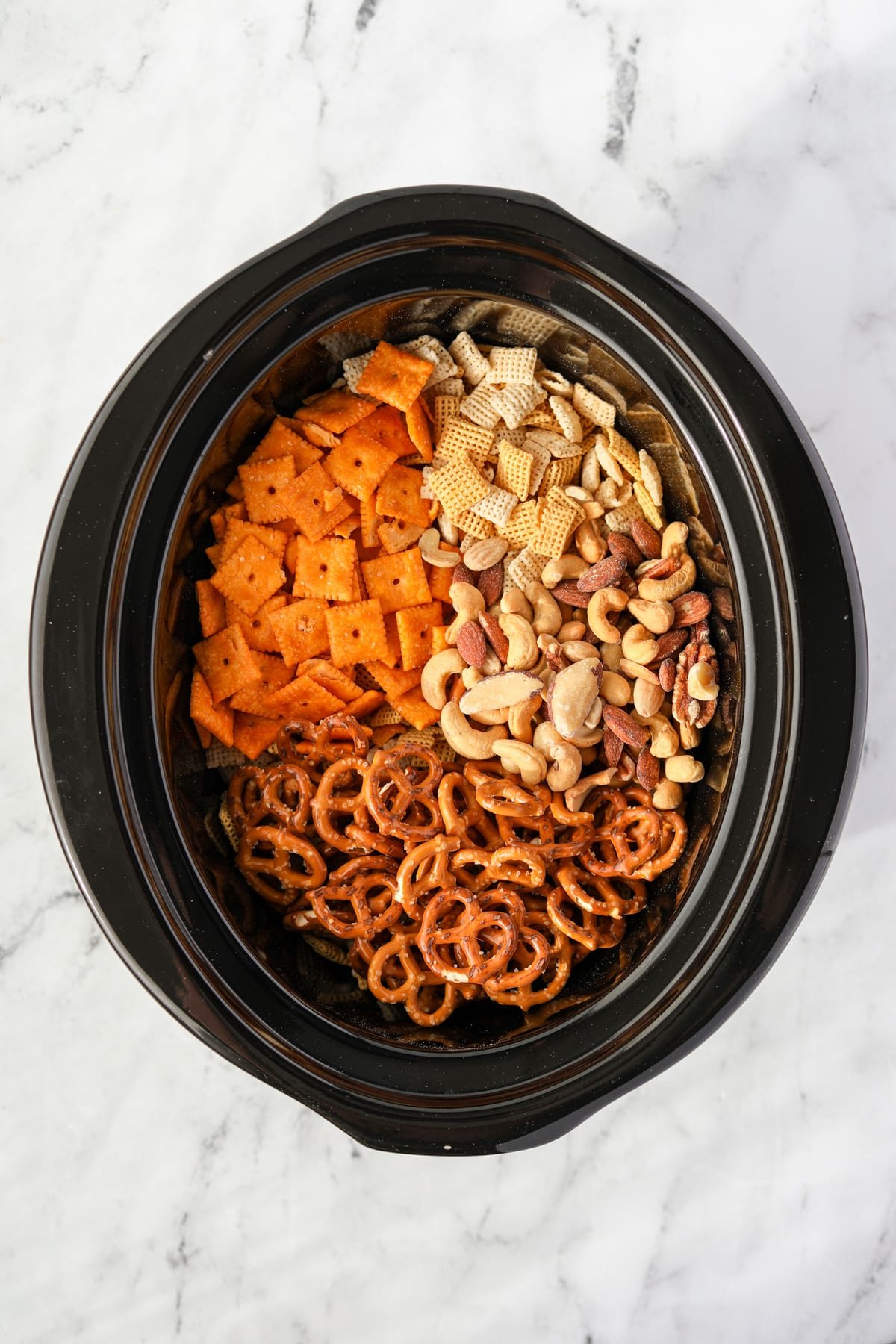 A slow cooker with chex cereal, crackers, pretzels, and mixed nuts inside/