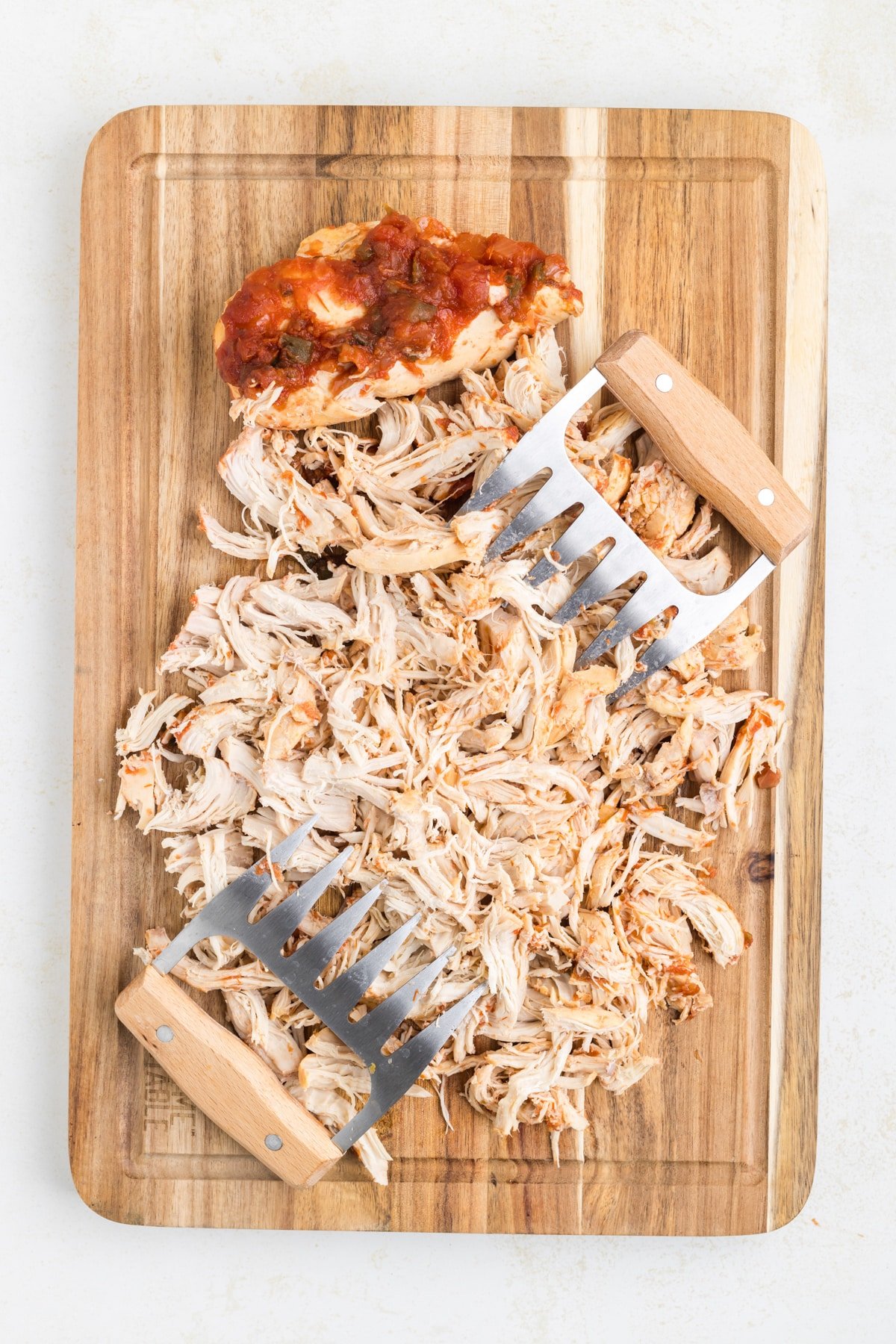 Using meat shredders to shred chicken breasts on a wooden cutting board.
