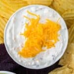 A bowl of creamy dip topped with cheese, with chips around the side.