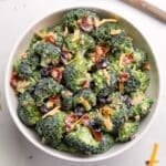 A white bowl filled with broccoli salad. It's garnished with cheddar cheese shreds and bacon.