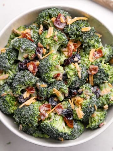 A white bowl filled with broccoli salad. It's garnished with cheddar cheese shreds and bacon.
