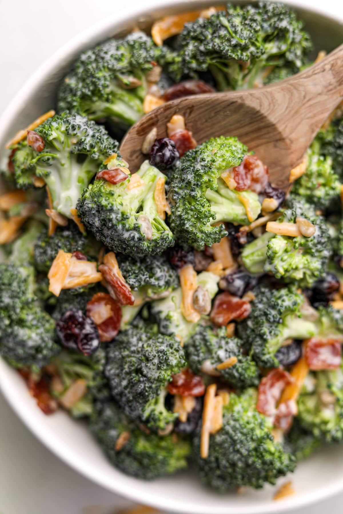 A wooden spoon taking a small portion from a large bowl of broccoli salad. It's garnished with bacon and cheddar cheese shreds.