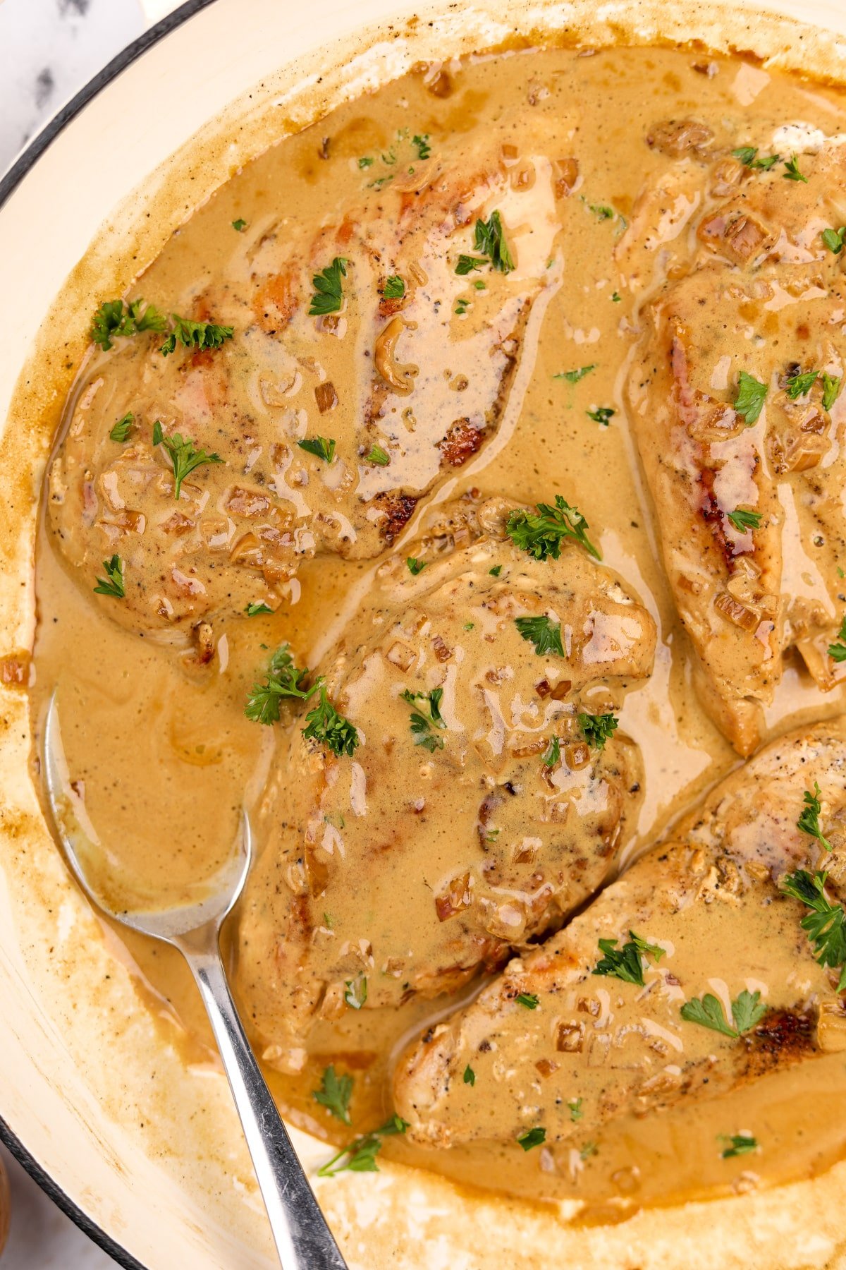 Four chicken breasts in a creamy pan sauce, with a spoon of sauce on the side.