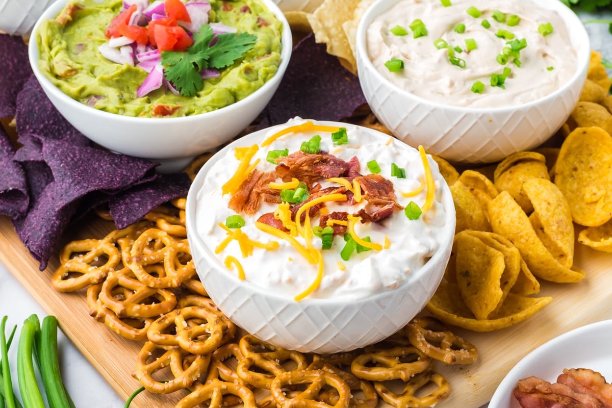 Three bowls of various dips, surrounded with blue corn tortilla chips, corn chips, and pretzels.