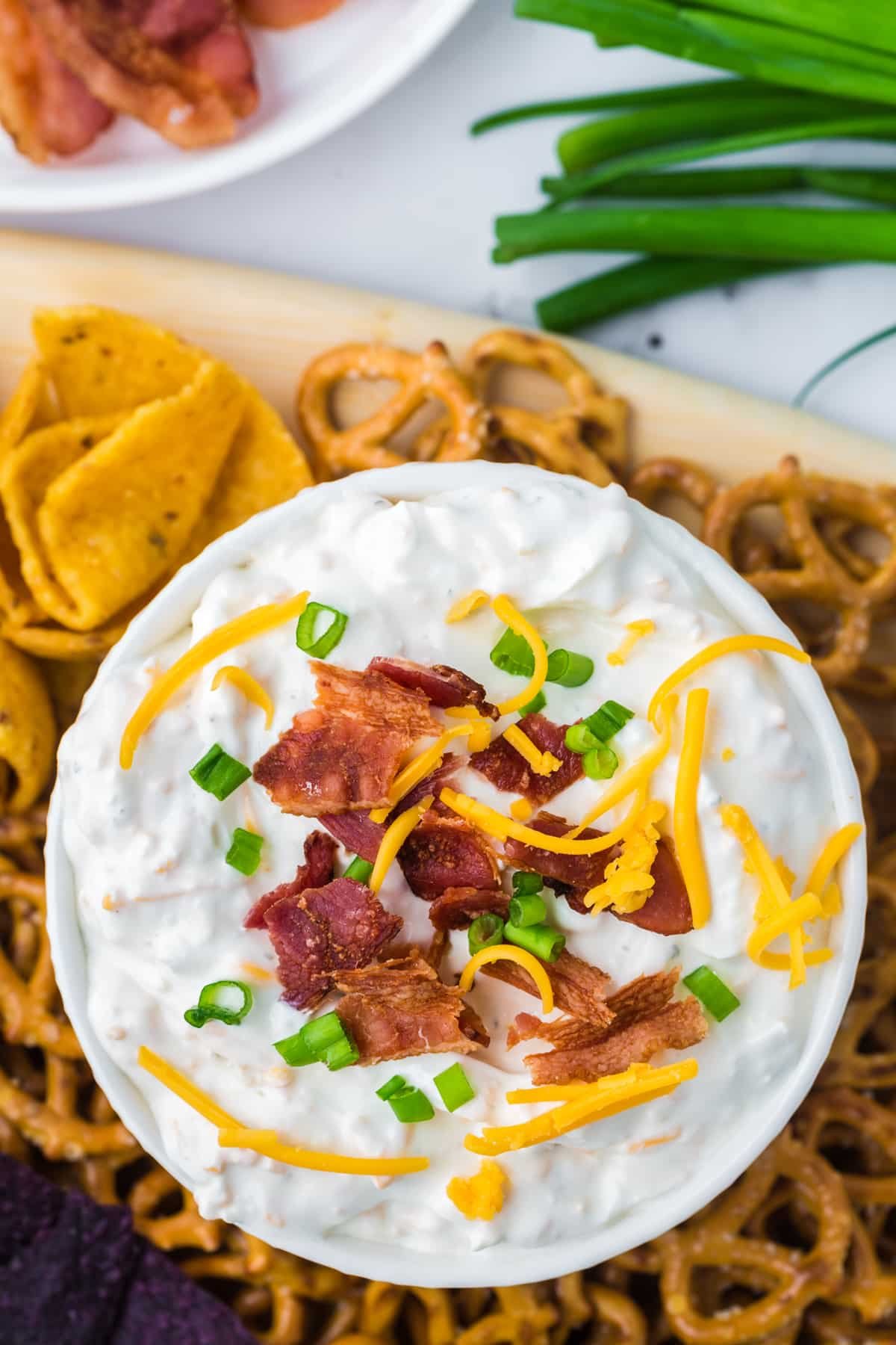 An overhead shot of a bowl of cream dip garnished with green onion, shredded cheese, and bacon. Surrounded by corn chips and pretzels.
