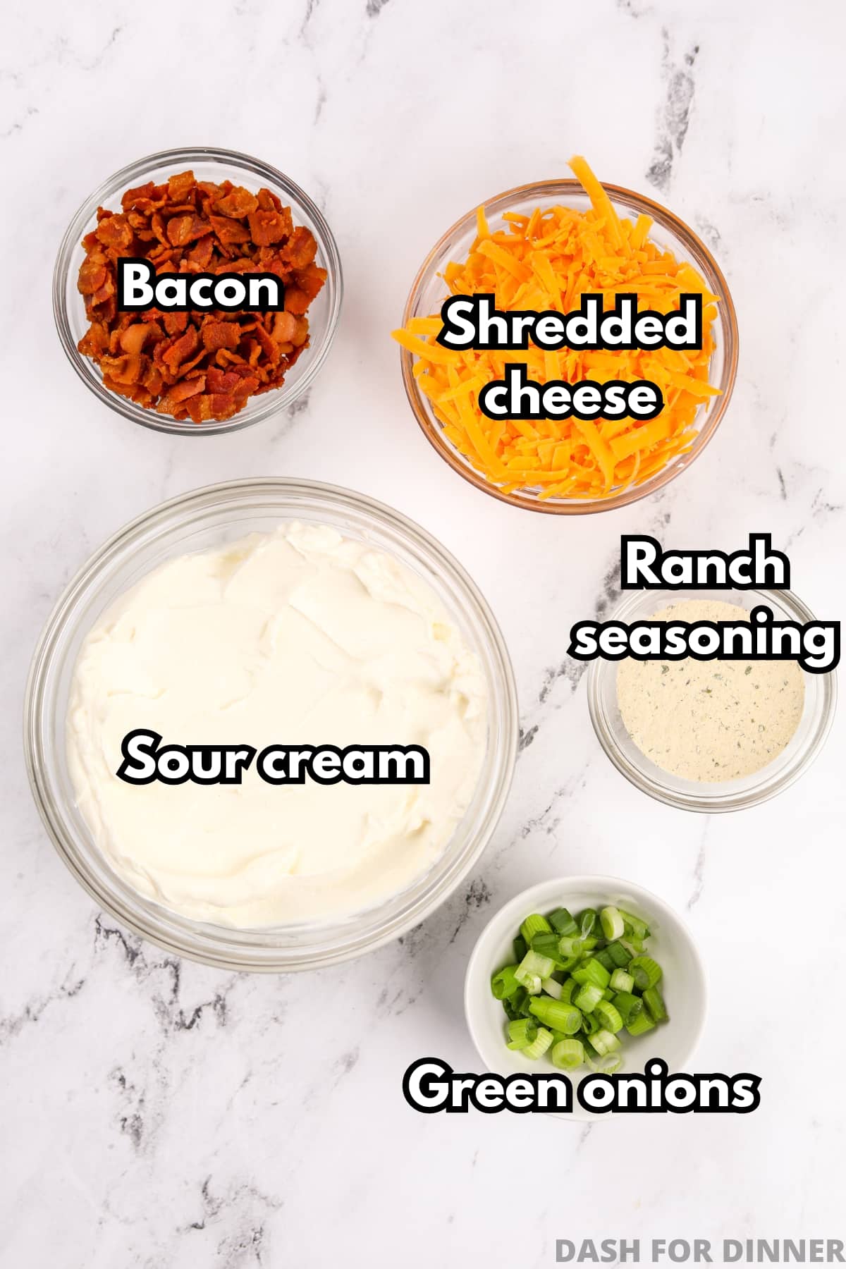 An overhead view of bowls of ingredients, including sour cream, shredded cheese, bacon, ranch seasoning, and green onions.