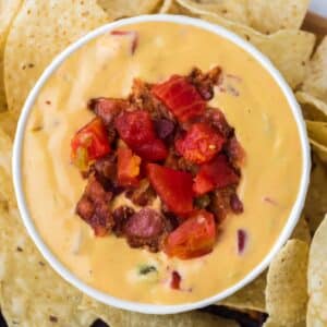 A white bowl filled with nacho cheese dip that's been garnished with bacon bits and tomato. Served with tortilla chips around the outside.