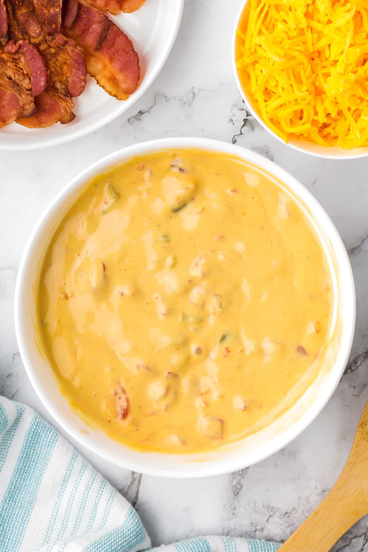 A large bowl of melted nacho cheese dip, with bowls of shredded cheese and bacon on the side.