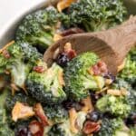 A wooden spoon scooping from a large bowl of broccoli salad garnished with bacon and cheese.