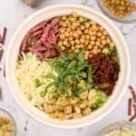 A bowl of chopped salad, featuring mozzarella cheese, artichokes, sun dried tomatoes, chickpeas, salami, and basil.